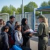 A group of people with backpacks and belongings waiting at a border checkpoint, speaking with uniformed officials.